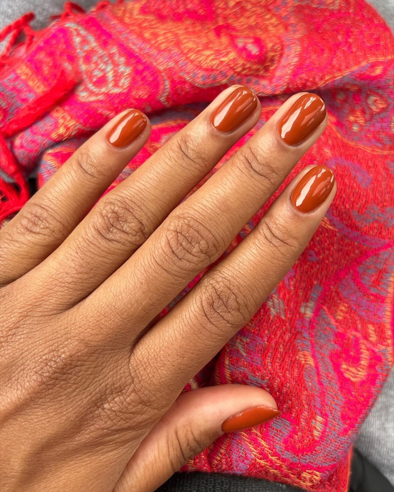 Glossy burnt caramel manicure on a deep tan hand over a pink paisley scarf