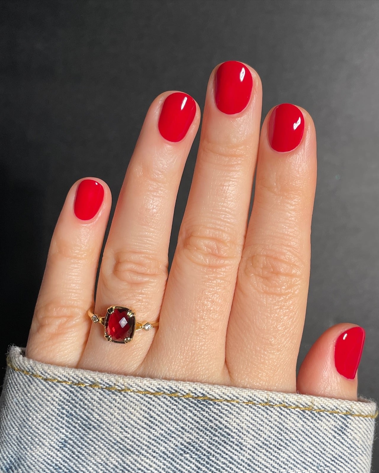 Bright true red short manicure on a fair hand with a garnet ring and denim jacket cuff